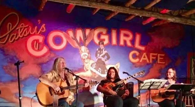 Nancy Scott's Songwriter's Circle - Three women performing live music with guitars at a cafe called 'Patsy's Cowgirl Cafe'. The background features a colorful mural with a cowgirl and vibrant neon-style lettering.