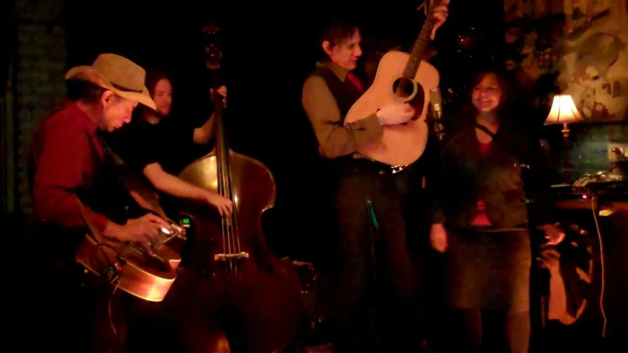 Allisong Smith and Greg Garing -  musicians performing on stage, playing guitar, upright bass, and keyboard, in a warm, cozy setting with a lamp and wall decorations in New York City.