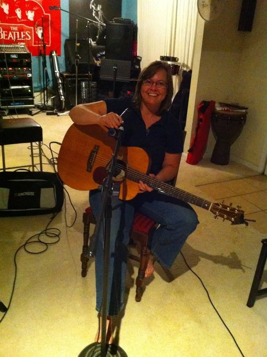 Allisong Smith Recording Session - Woman sitting on a wooden chair playing an acoustic guitar, smiling, in a music studio with equipment and posters in the background.