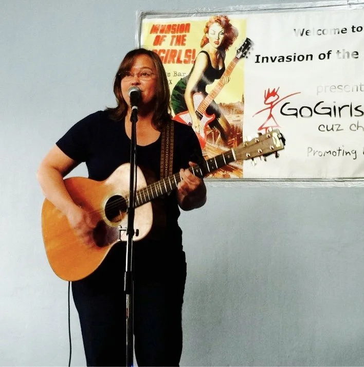 Allisong Smith - A woman singing and playing an acoustic guitar on a stage with a microphone. Behind her is a poster of a girl with red hair holding a guitar and a whiteboard with text that reads 'GoGirls Music' and other words.