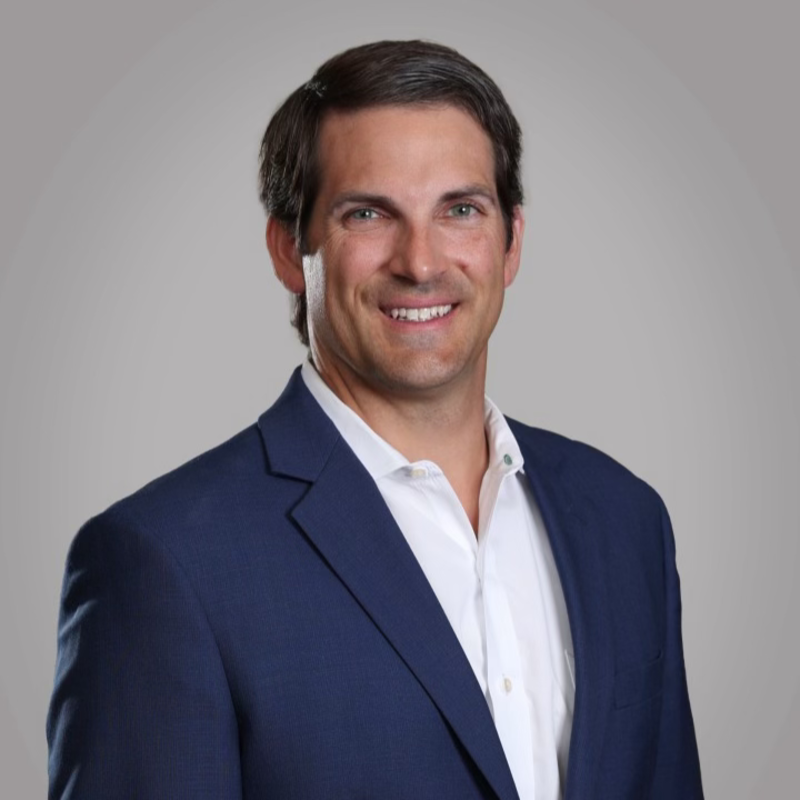 A smiling man wearing a navy suit and white shirt against a plain gray background.