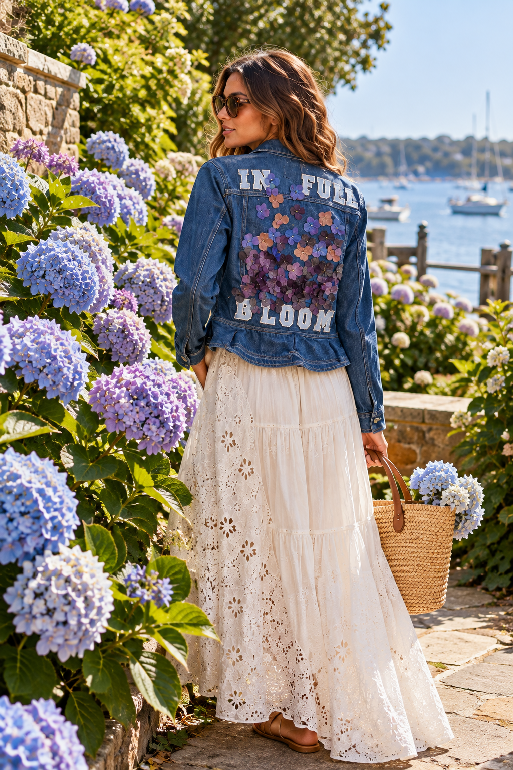 A woman standing among blue and purple hydrangeas by a waterfront, wearing sunglasses, a denim jacket with floral applique and the words 'IN FULL BLOOM', a white lace skirt, and holding a straw tote bag with hydrangeas inside.