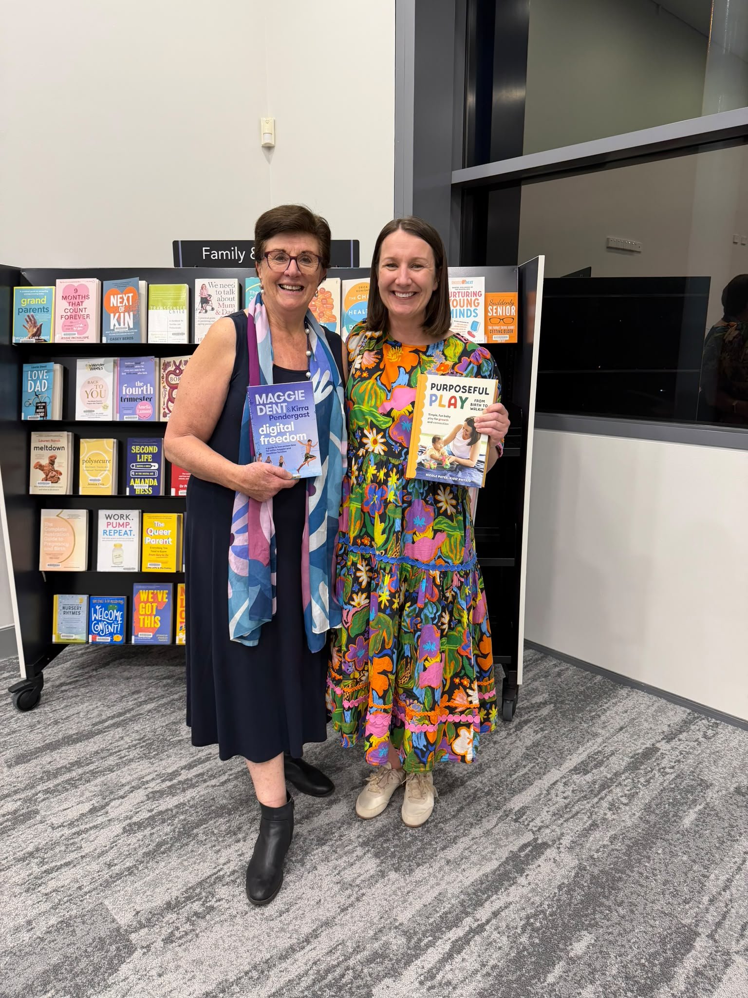 Two women standing side by side in front of a book display at a library or bookstore, smiling at the camera. The woman on the left is wearing a black dress, black boots, glasses, and a colorful scarf, holding a book titled 'Digital Freedom'. The woman on the right is wearing a vibrant, floral-patterned dress and beige sneakers, holding a book titled 'Purposeful Play'.