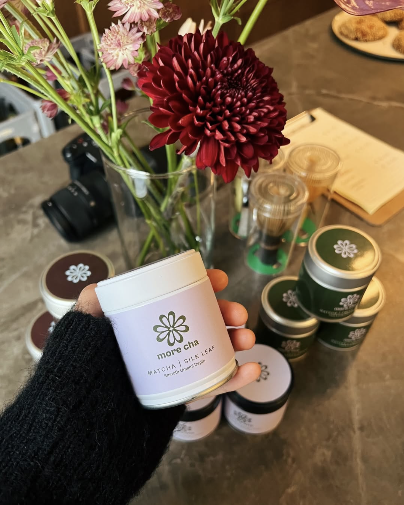 A hand holding a container of 'more cha' matcha silk leaf with a pink and white label, in front of a table with arranged matcha containers, a vase of pink and red flowers, a camera, and a notebook.