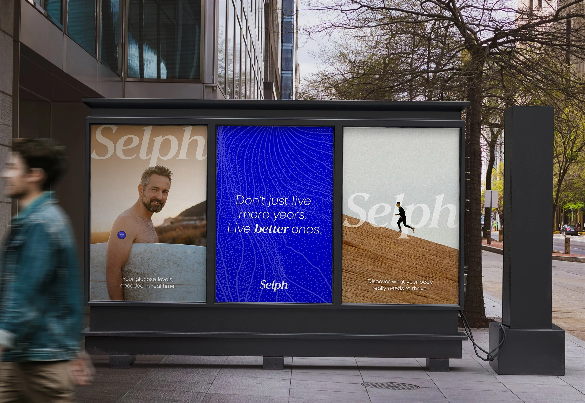 A large outdoor advertisement display with multiple panels on a city sidewalk. The left panel shows a man with a tattoo, and the text "Your glucose levels, decoded in real time." The middle panel features a blue patterned background with white text: "Don't just live more years, Live better ones," and the brand name "Selph." The right panel displays a person running on a dirt trail with the text "Discover what your body really needs to thrive." The scene is set against a backdrop of a modern building with glass windows and leafless trees, with two blurred pedestrians walking past.