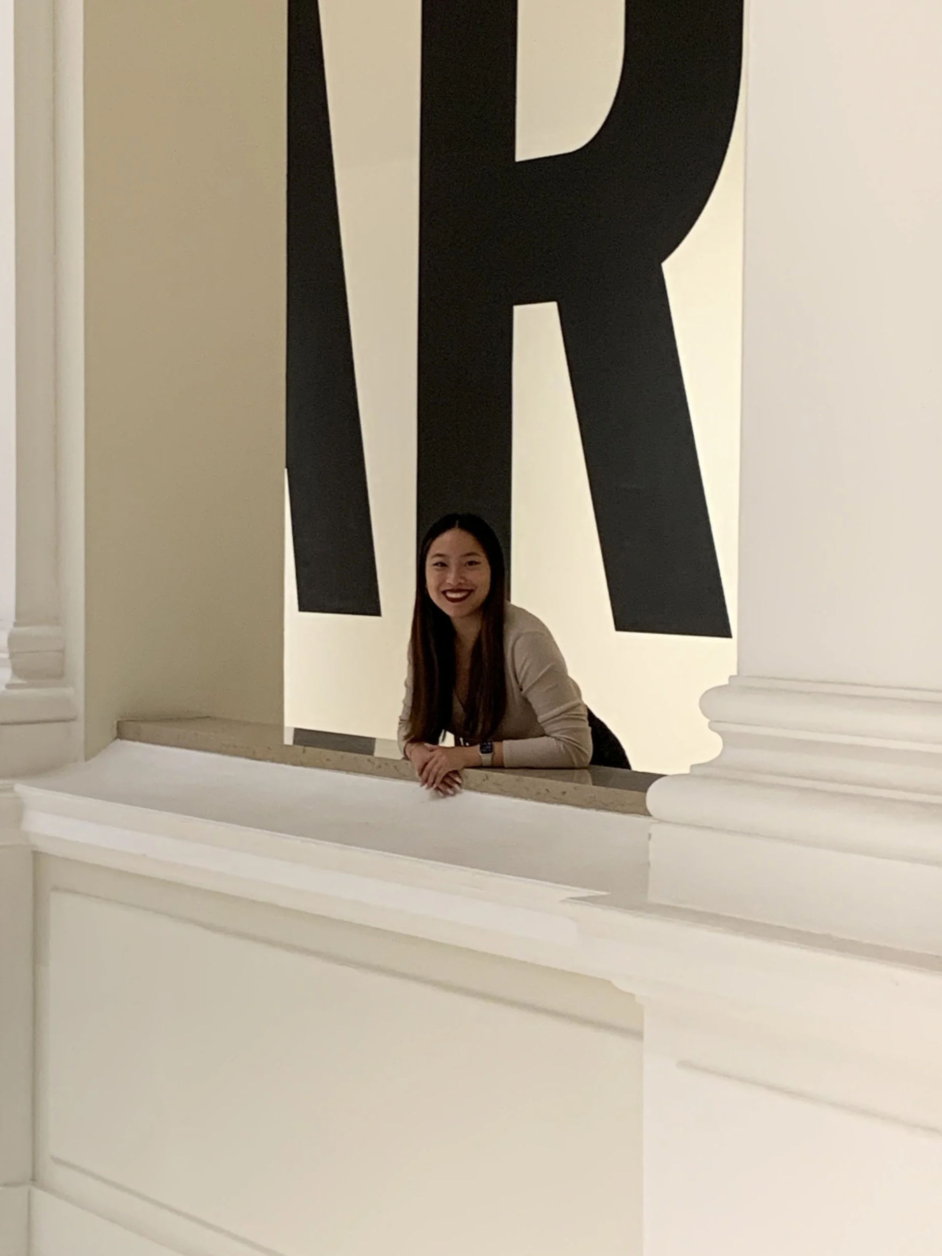A woman with long dark hair and dark lipstick sitting on a white ledge in front of a large black and white abstract art piece. The art piece features large bold black shapes on a white background.