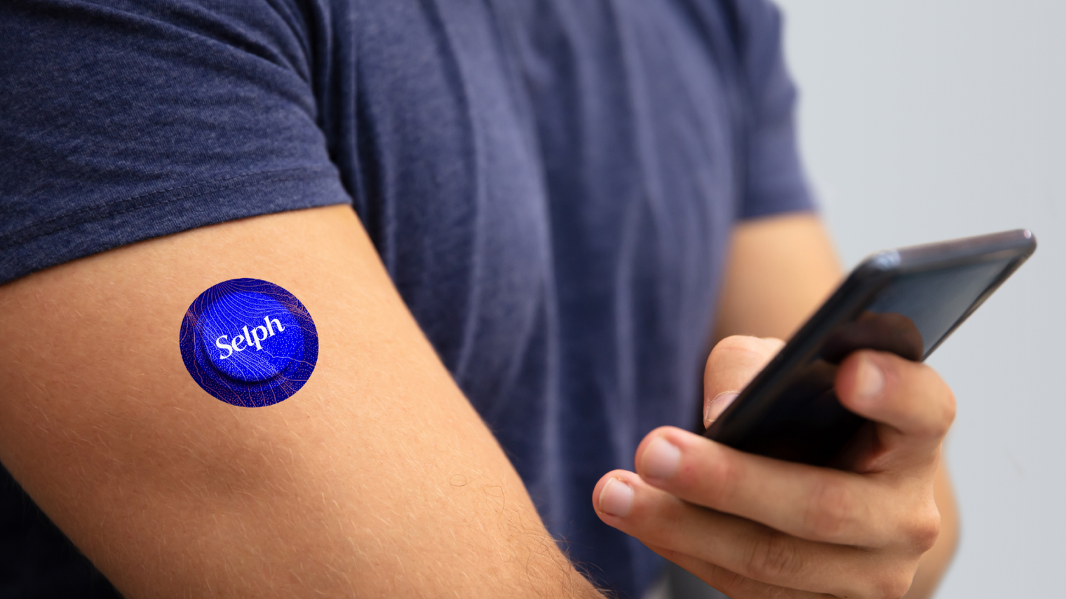 A person's upper arm with a blue circular COVID-19 vaccine sticker that says 'Selph' on it, while the person looks at a smartphone in their hand.