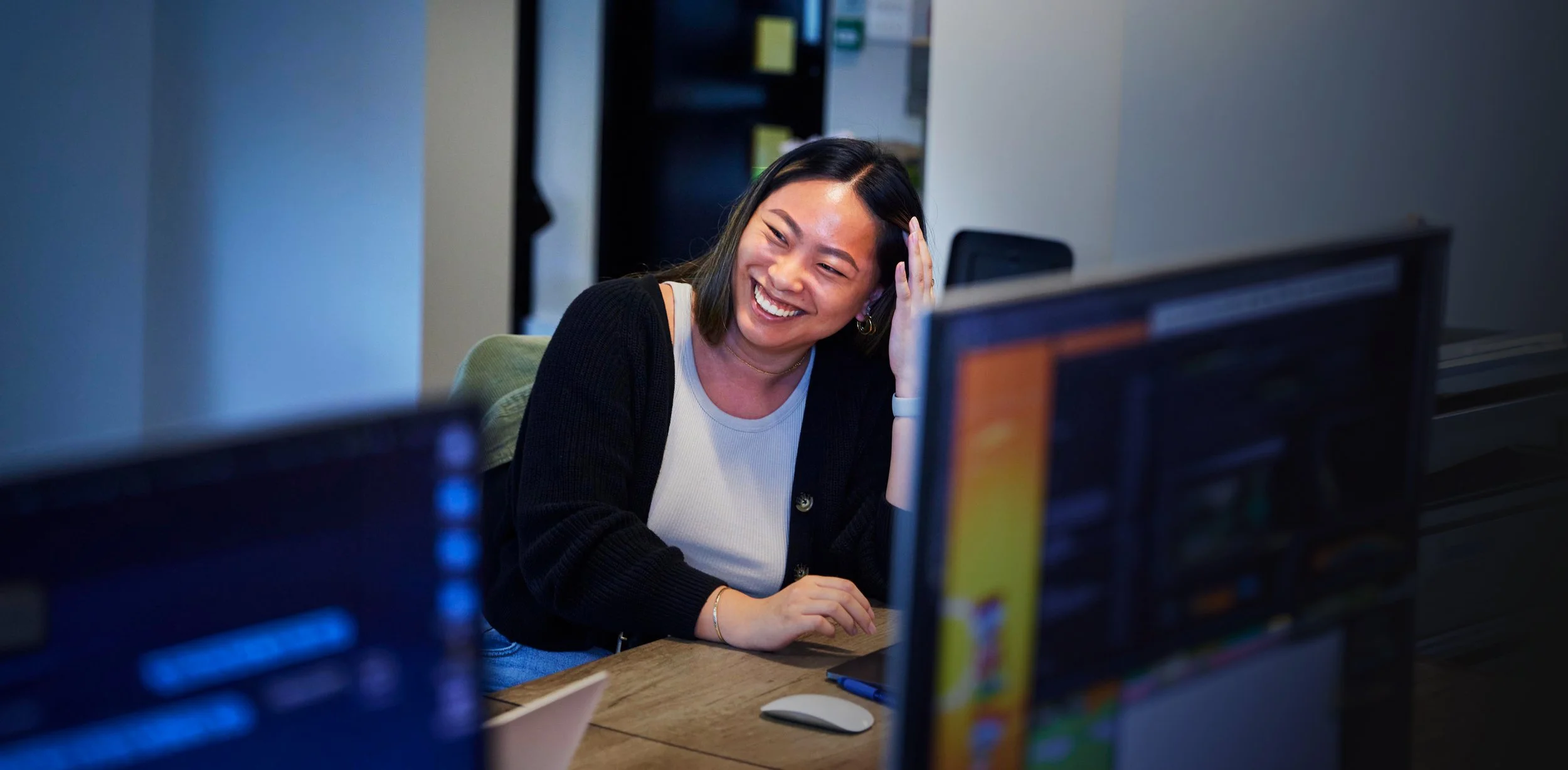 A woman with black hair smiling and touching her head while working at her desk in an office, with multiple computer monitors and office shelves in the background.