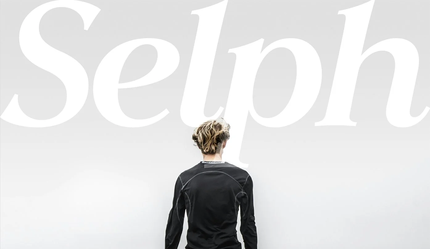 A person with short, wavy blonde hair wearing a black jacket, facing a large, light gray wall with the word 'Sep' partially visible.