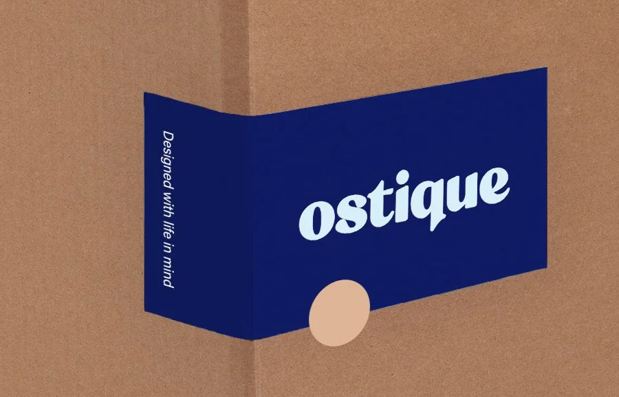 A navy blue fold-out booklet with the word "ostique" in large white letters and the phrase "Designed with life in mind" on the side, placed on a brown background.