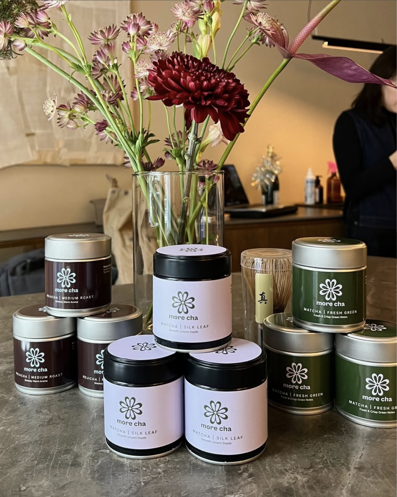 A clear glass vase with pink and maroon flowers, including a large maroon dahlia, placed on a counter. Several containers of matcha tea products labeled, including 'more cha' and 'Matcha | Silk Leaf' and 'Matcha | Fresh Green', are arranged in front of the vase. In the background, a person in a black top and a wall with a light-colored curtain can be seen.
