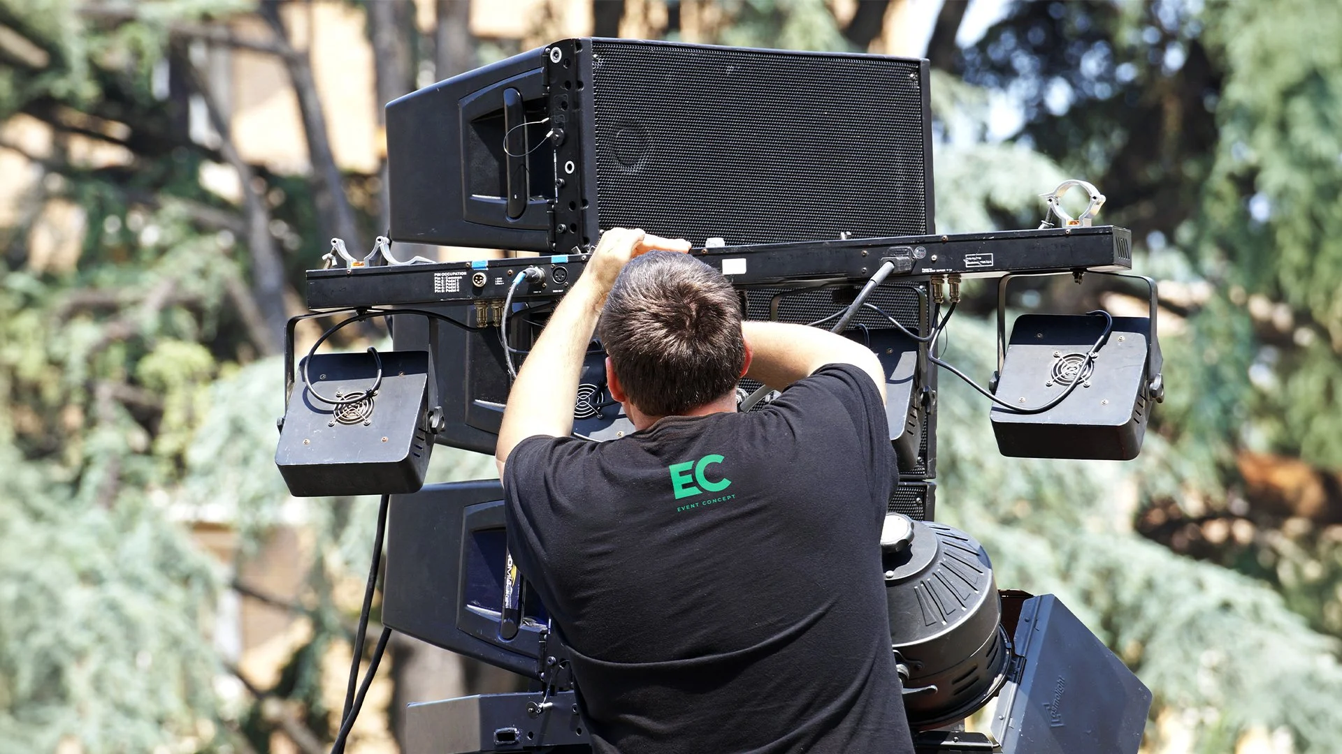A cameraman operating a large professional video camera outdoors during daytime, with trees in the background.