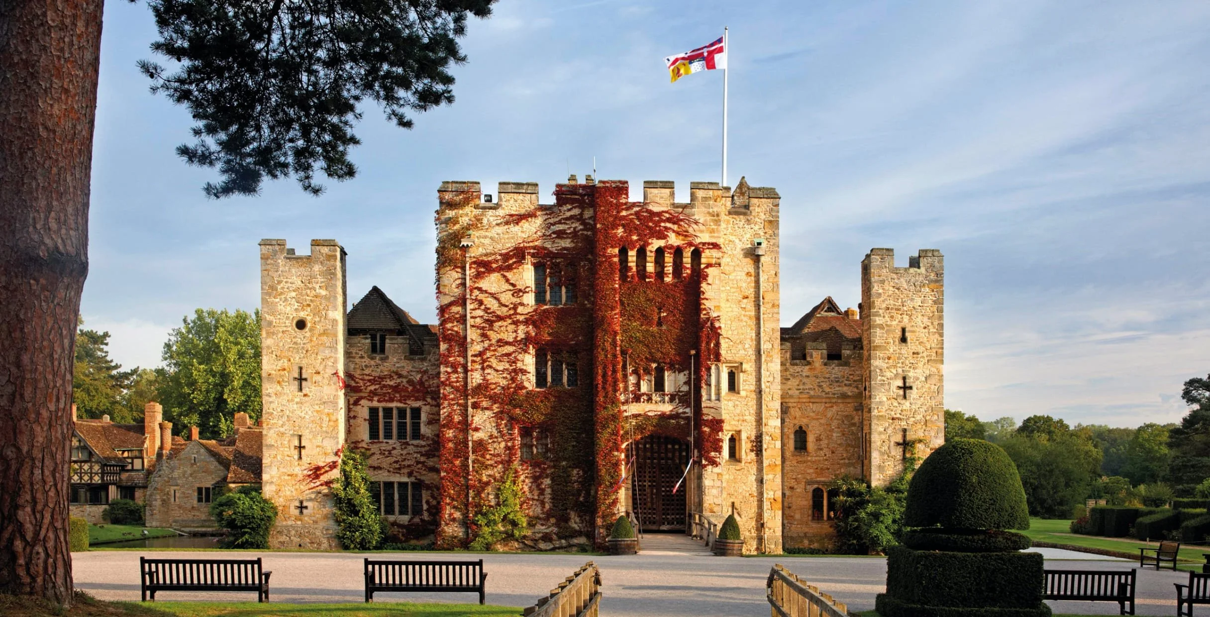 A historic stone castle with ivy-covered walls, part of a park with benches, lawns, and trees, under a partly cloudy sky.
