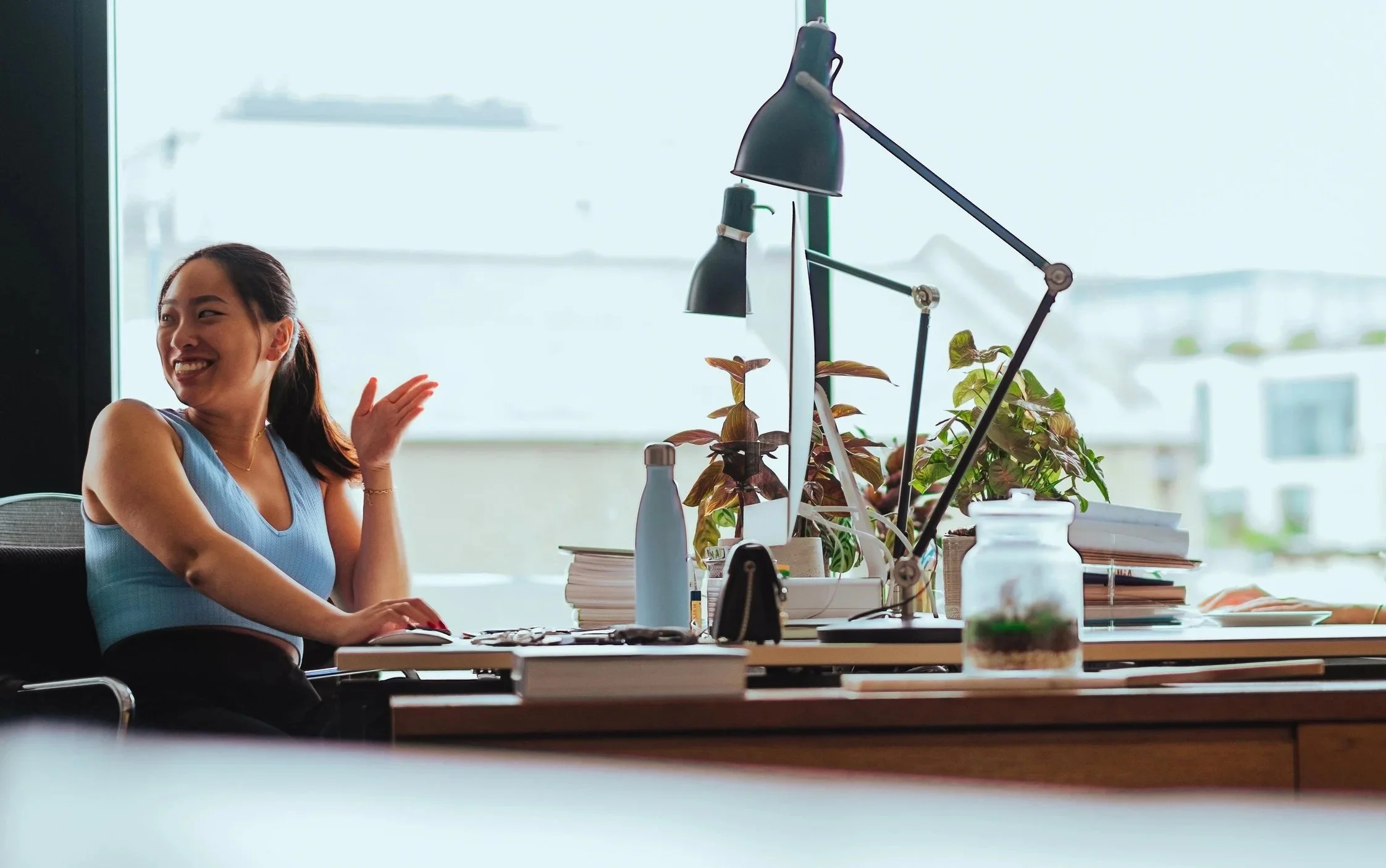A woman in a blue sleeveless top smiling and waving at her office desk, which has plants, books, and various objects.
