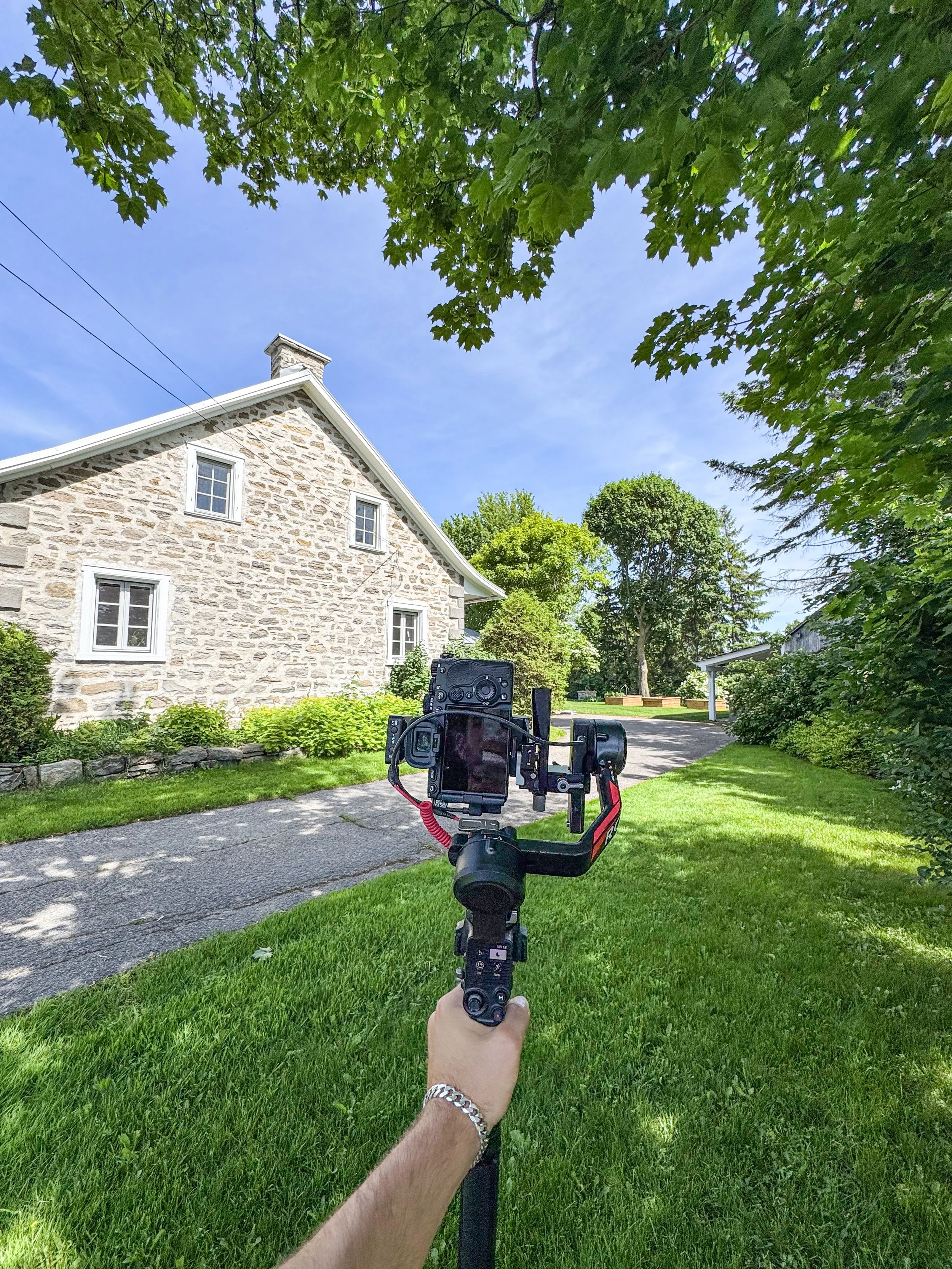 Prise de vue d'une caméra sur trépied pointant vers une maison en pierre avec un jardin verdoyant et des arbres, sous un ciel bleu avec quelques nuages, photo capturée par la personne portant une montre en argent et un bracelet en chaîne.