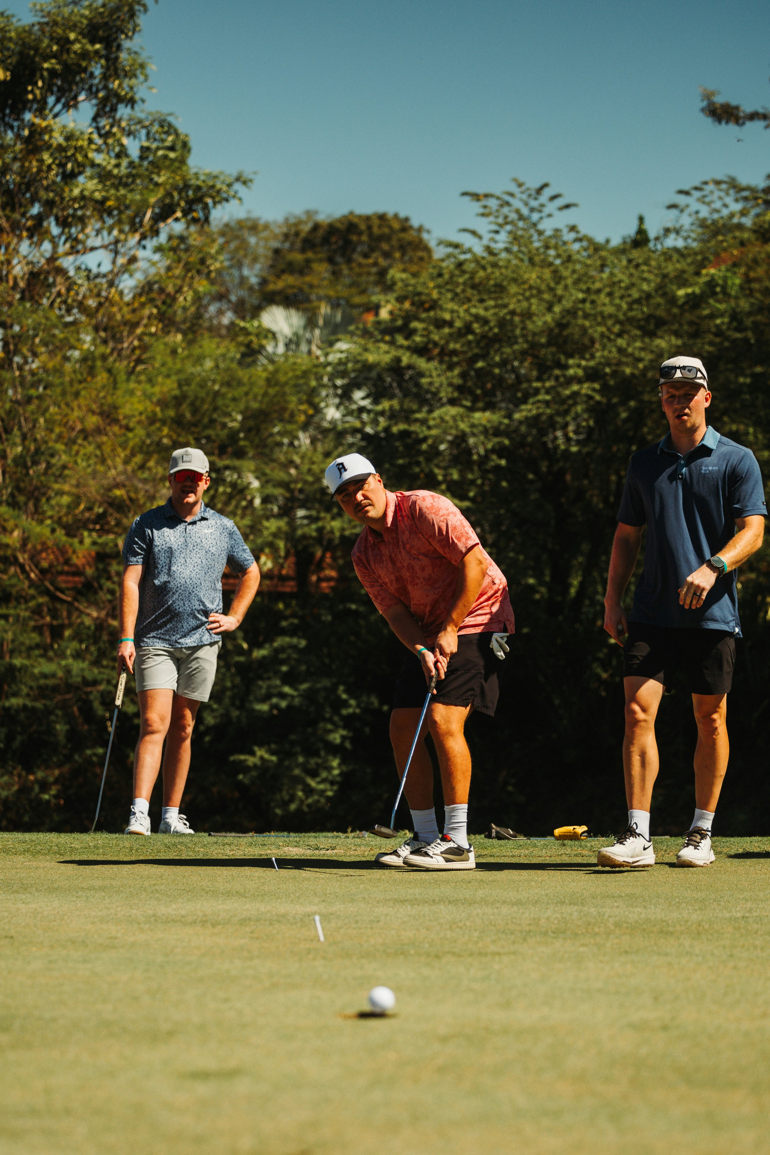 Four men on a golf course playing golf, with one preparing to putt and the others watching, surrounded by trees under a clear sky.