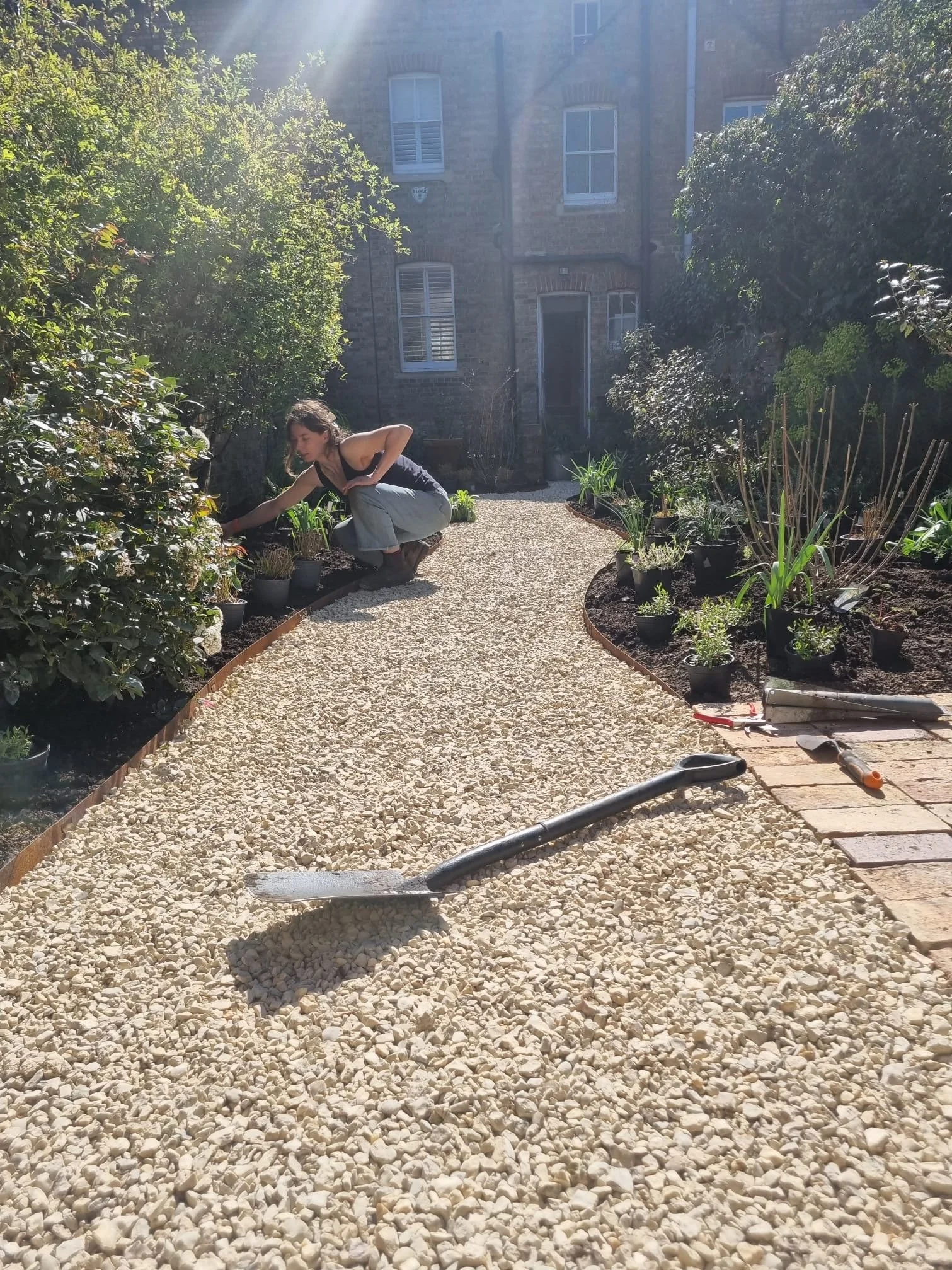 A woman is gardening in a backyard, crouching on a gravel path surrounded by plants and potted greenery, with a brick-built house in the background and gardening tools on the ground.