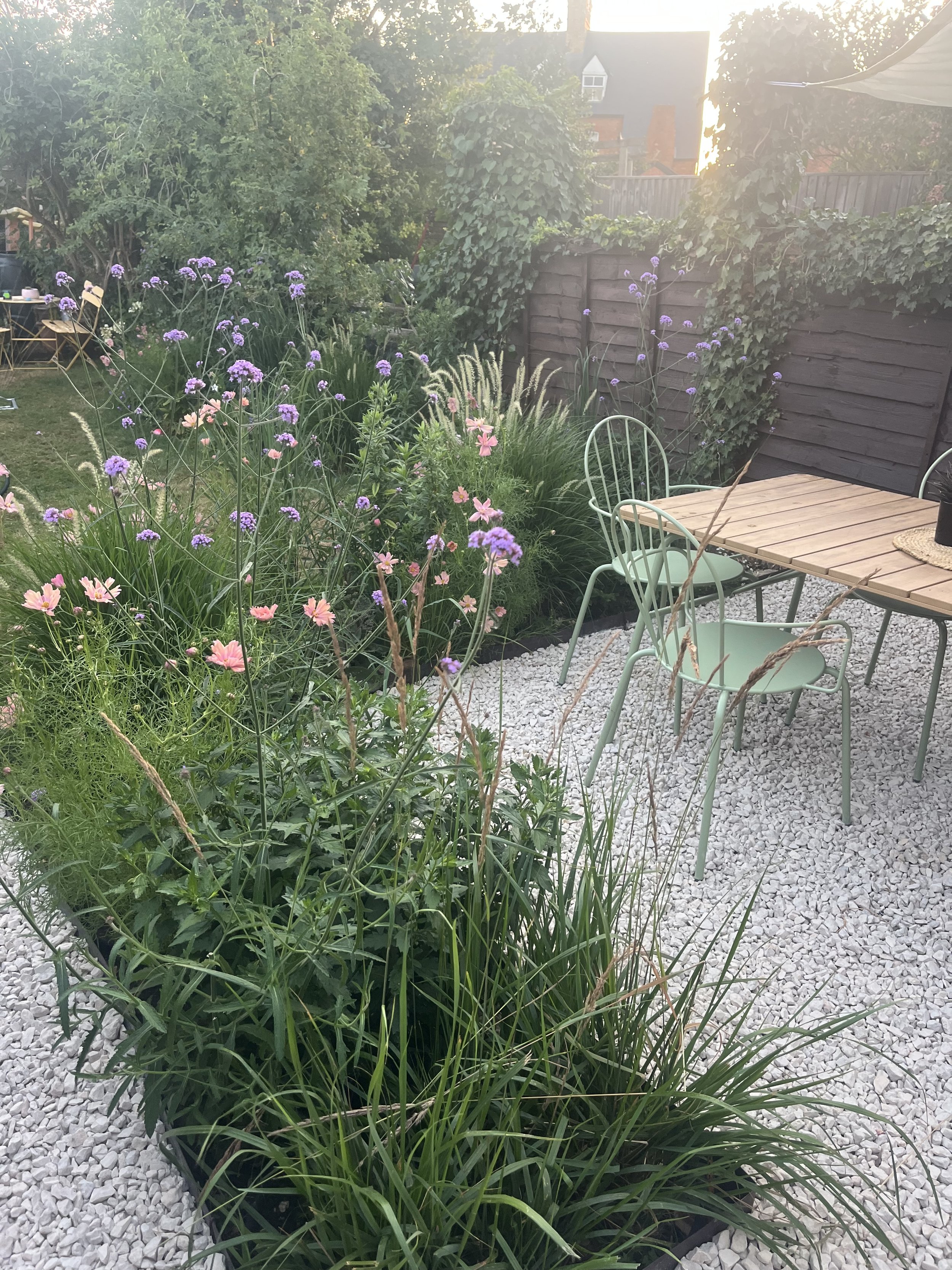 A garden patio with green chairs and a wooden table surrounded by flowering plants, a wooden fence, and trees in the background with sunlight.