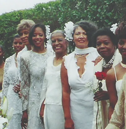 Group of women dressed in white, standing outdoors, some holding roses, smiling, with green hedge background.
