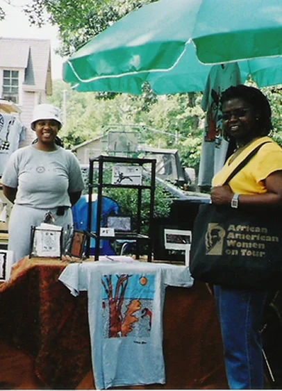 Two women standing behind a table at an outdoor market under a palm leaf pattern umbrella, with books and artwork on display.