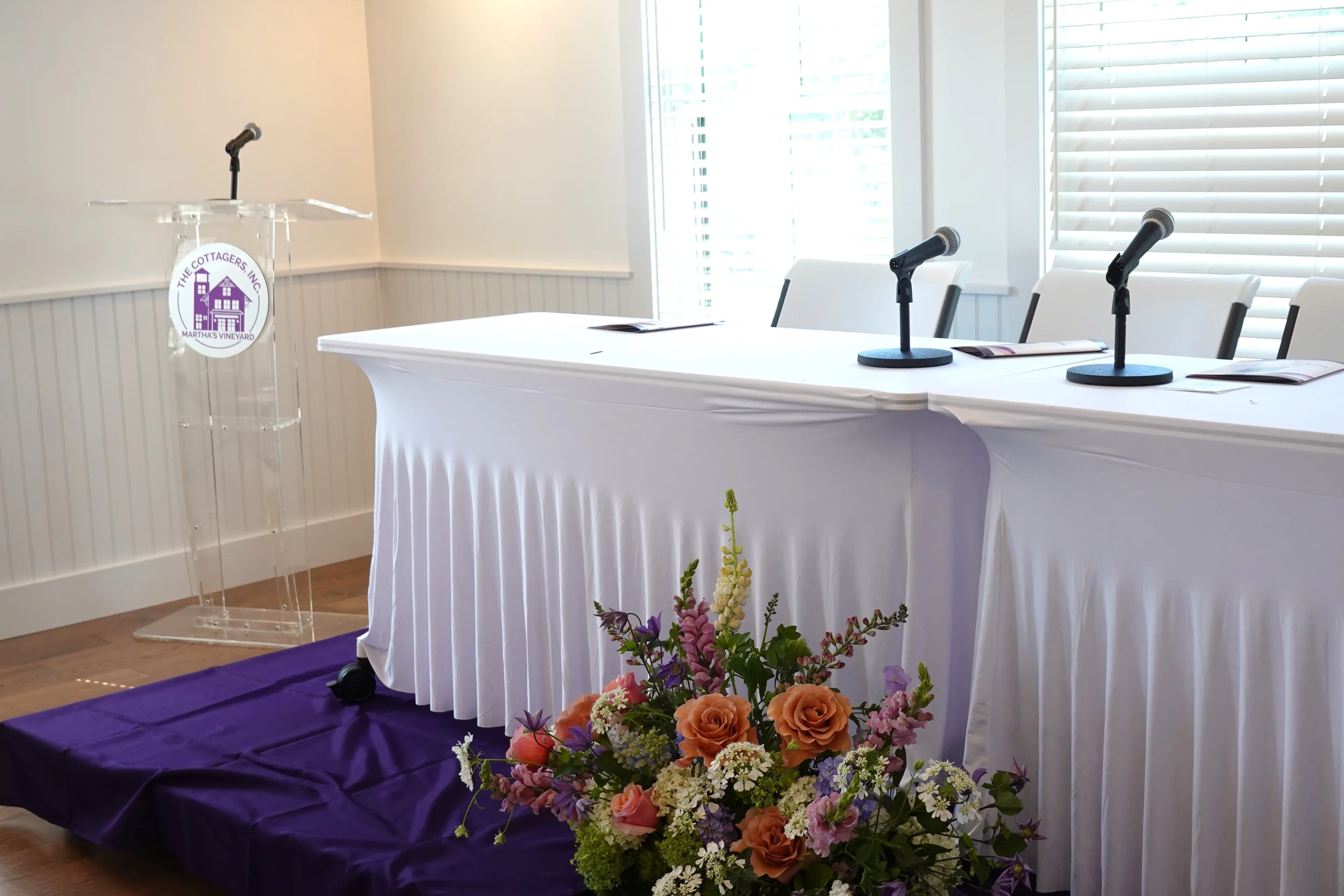 An empty panel discussion or presentation setup with three microphones on a table, a clear podium with logo, and a floral arrangement in front, in a brightly lit room with large windows and white walls.