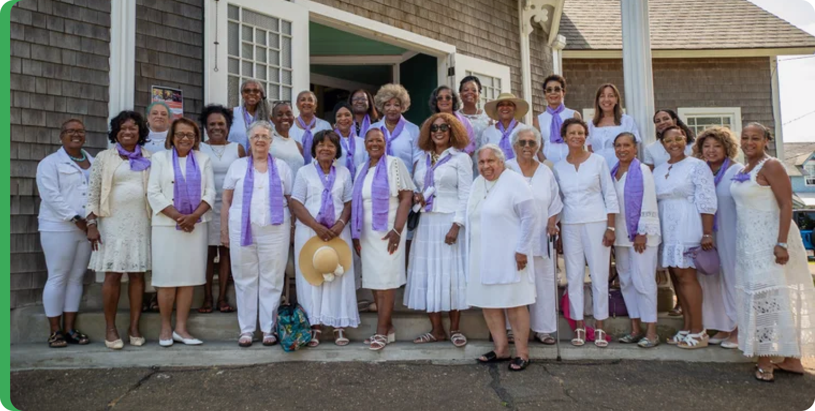 Group of women standing on the porch steps of a house, dressed in white, with many wearing purple scarves around their necks, smiling for the camera on a sunny day.