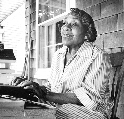 An elderly woman sitting outdoors at a wooden deck, with a window behind her, wearing a striped shirt and looking thoughtfully into the distance.