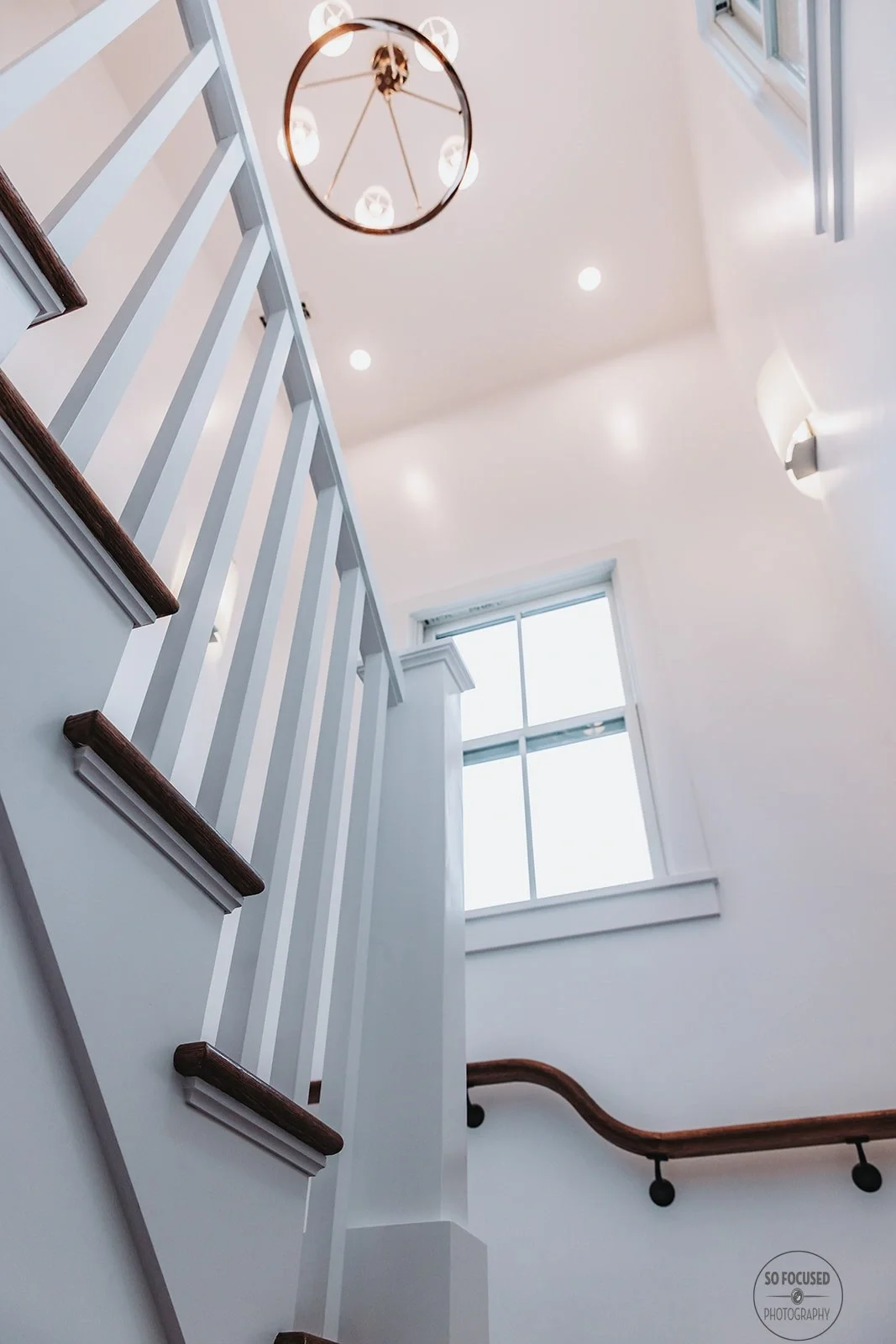 Interior of a modern house with white walls, a tall window, a wooden staircase with white spindles, a curved wooden handrail, and a contemporary circular chandelier hanging from the ceiling.