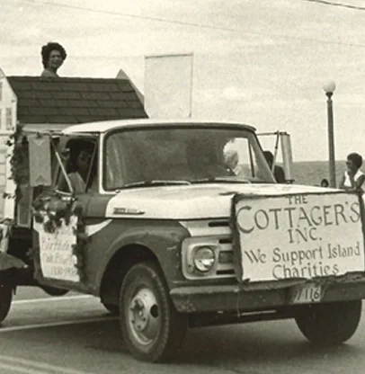 Vintage truck with a sign for The Cottagers Inc. supporting island charities, parked on a street with people and a lamppost nearby.
