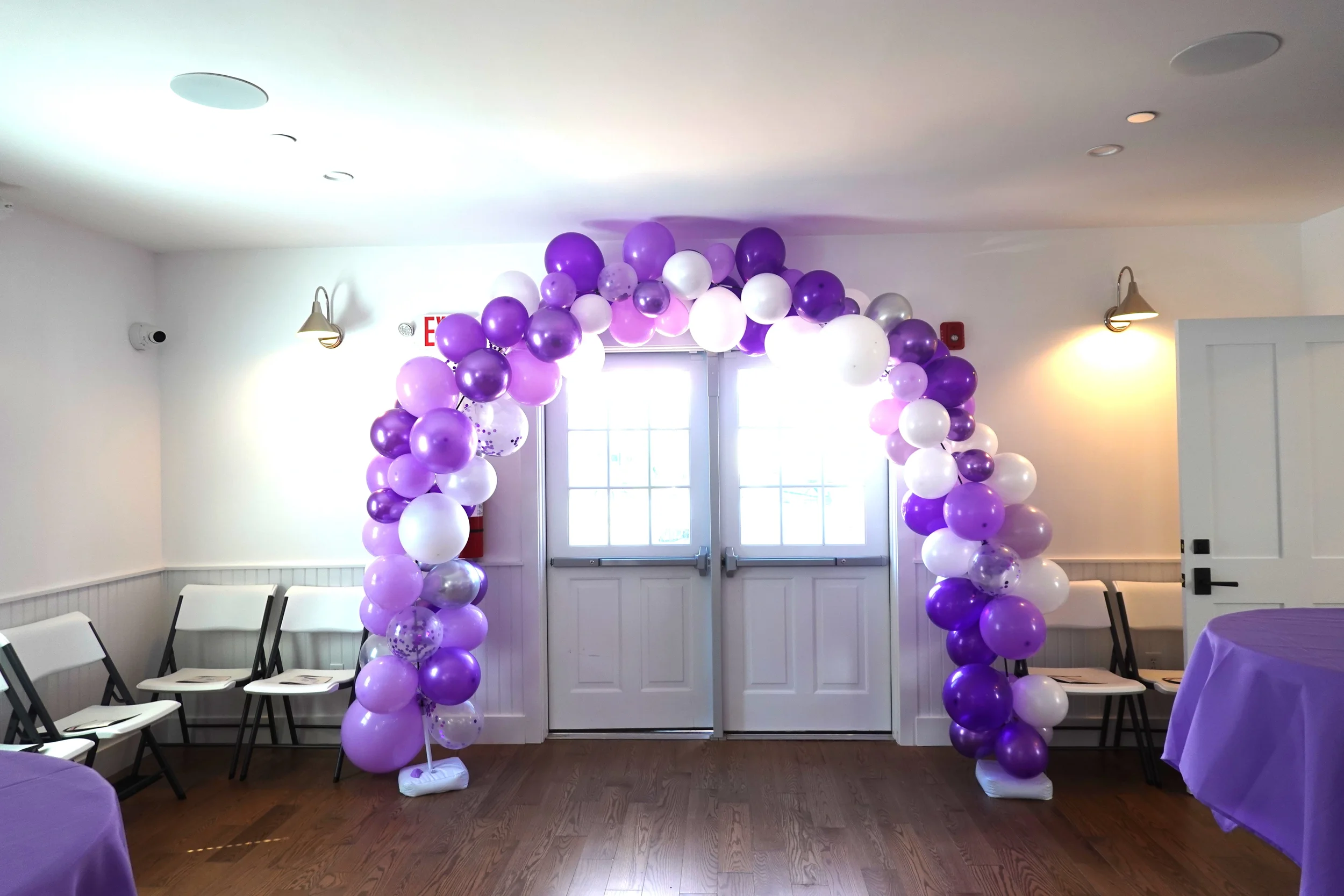 Indoor event space decorated with a balloon arch in purple, white, and lavender, with chairs arranged on either side and tables covered with purple tablecloths.
