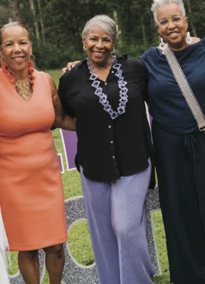 Three smiling women standing outdoors, dressed in colorful attire, with greenery in the background.