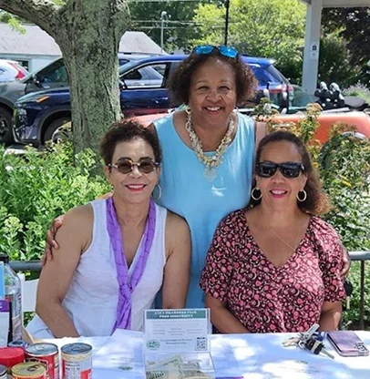 Three women smiling at an outdoor table with food and drinks, two women wearing sunglasses, one in a Purple scarf and white top, the other in a pink floral top, and the woman in the middle standing behind them in a blue top with a necklace.