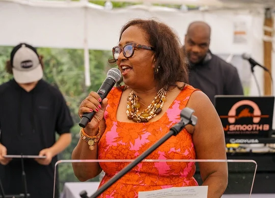 Woman speaking into a microphone at an outdoor event, wearing glasses, a bright orange dress with pink floral pattern, and accessorized with jewelry, with two men in the background, one using a DJ equipment.