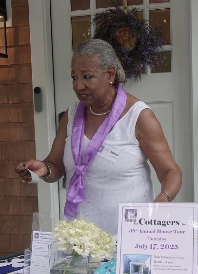An elderly woman with gray hair wearing a white sleeveless dress and a purple scarf, standing at a table with informational materials at an outdoor event.