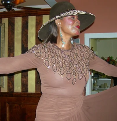 Woman wearing a large hat, elegant dress, and jewelry, standing with arms outstretched indoors.