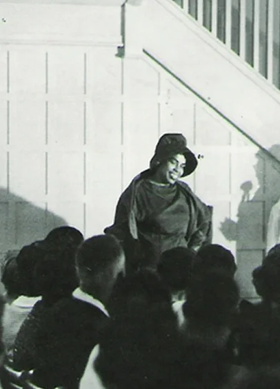 A woman standing and speaking to a group of children indoors, with a staircase and railing in the background.