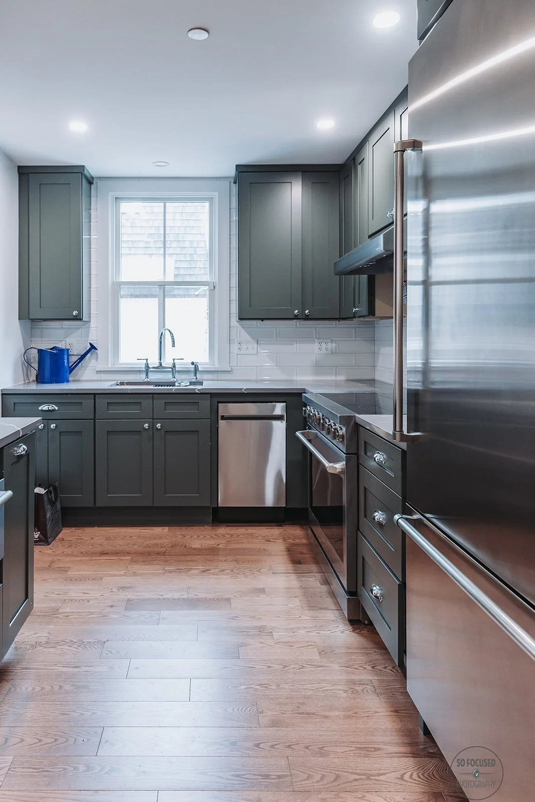 Kitchen with gray cabinets, stainless steel appliances, wooden floor, white tiled backsplash, window above the sink, and ceiling lights.