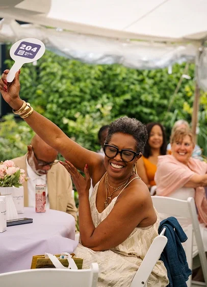 Woman wearing glasses and a white dress, smiling and holding a sign, at a social gathering under a tent with other smiling women in the background.