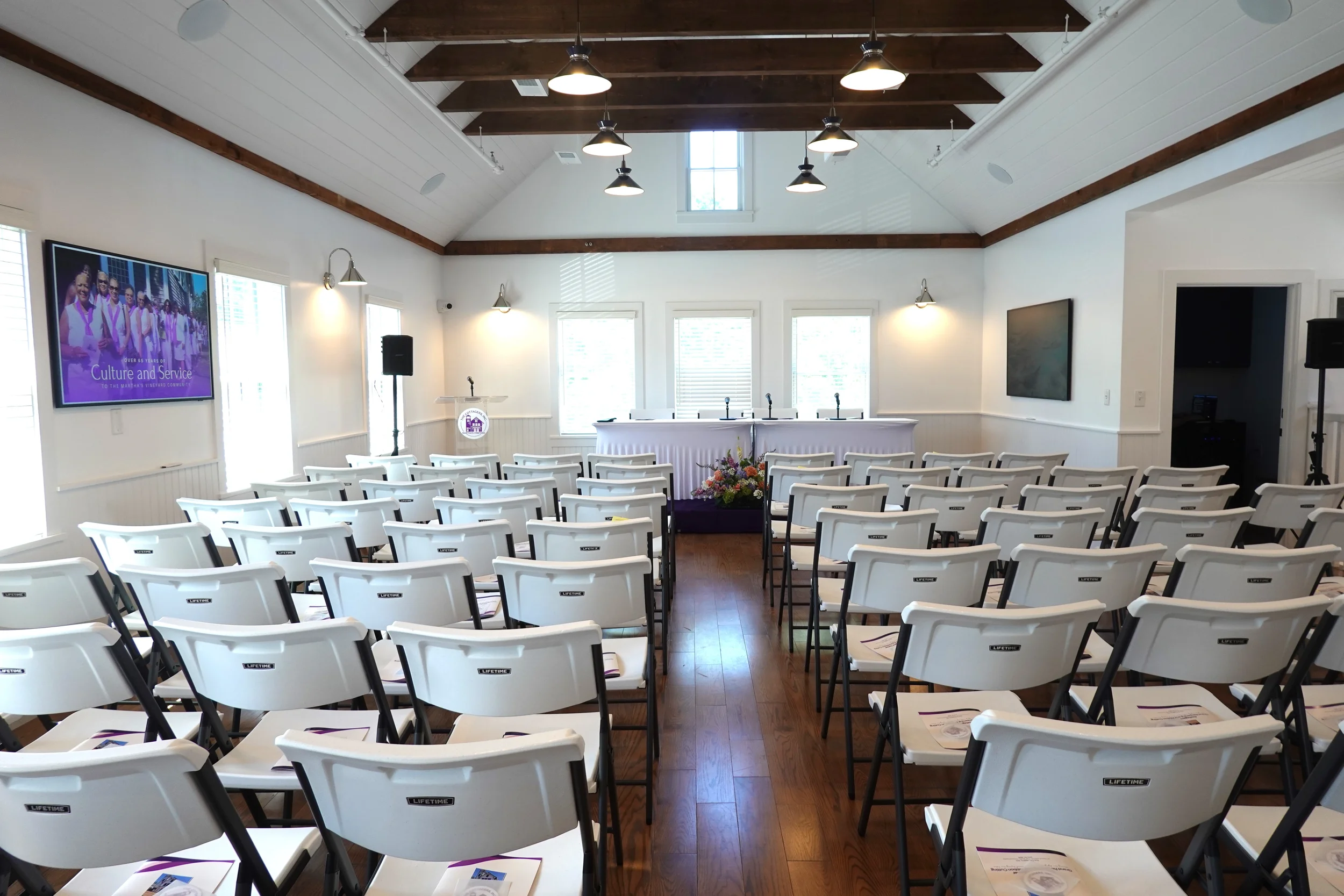 A spacious room set up for a presentation or ceremony, with rows of white chairs facing a head table with microphones. There are large windows, a screen displaying a slide titled 'Culture and Service,' and ceiling lights, with wood beams on the ceili