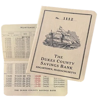 A booklet labeled 'The Dukes County Savings Bank' with an illustration of a sailing ship on the cover, placed beside a printed mortgage statement or bank document.