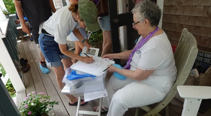 A woman sitting on a porch chair at a table, signing documents, while a young girl and other people stand nearby. The woman appears to be engaged in paperwork with others around.