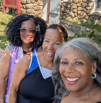 Three women smiling outdoors in front of a stone building and grassy area on a sunny day.