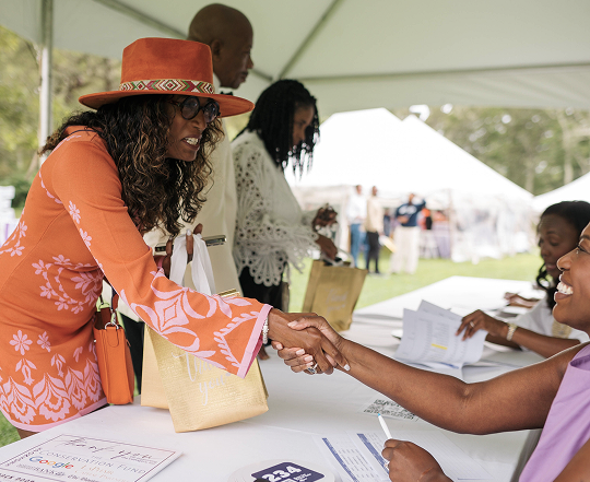 Woman in orange blouse and hat shaking hands with a seated woman at an outdoor event under a white tent.