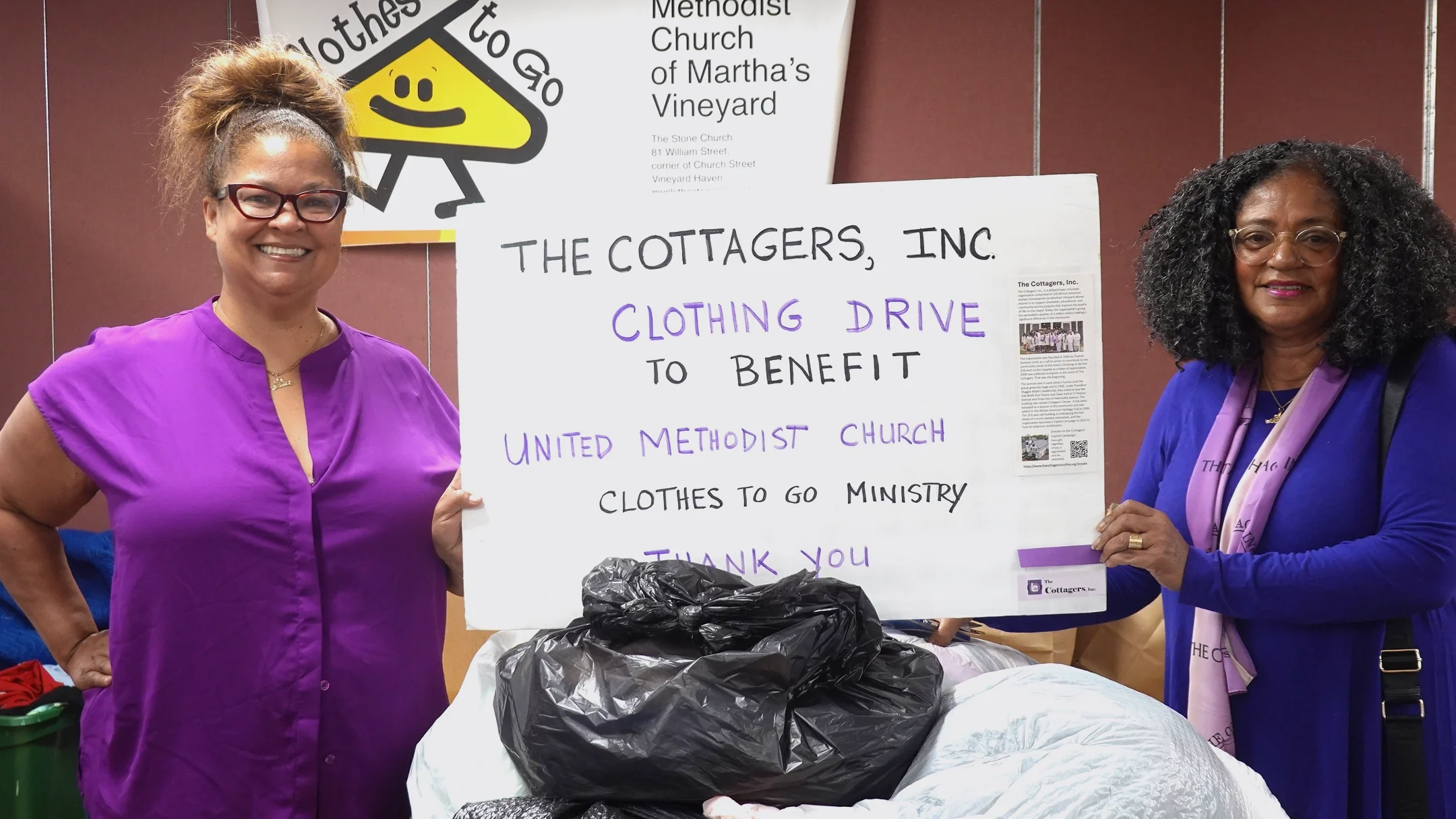Two women standing indoors, smiling, holding a large sign that reads 'The Cottagers, Inc. Clothing Drive to Benefit United Methodist Church Clothes to Go Ministry. Thank You'. The woman on the left is wearing a purple top, glasses, and has her hair in a high puff. The woman on the right is wearing a blue top, glasses, and a pink scarf, holding a purple card.