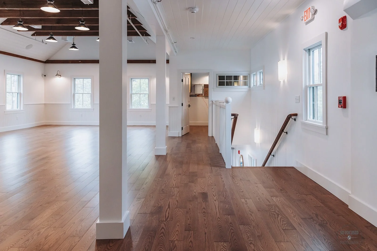 Empty interior of a modern house with white walls and wooden floors, featuring multiple windows, a staircase, and ceiling lights.