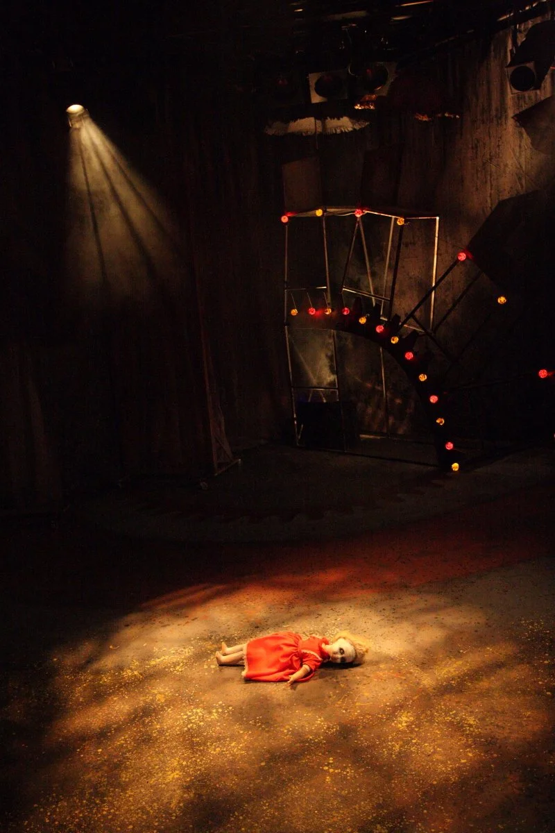 A puppet girl with blonde hair in a red dress lying on the stage floor with a spotlight shining down on her, dark stage with a set piece in the background resembling part of funfair big wheel.