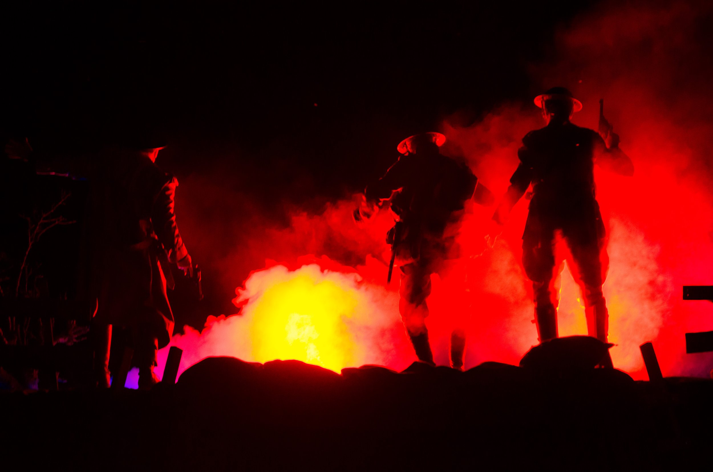 Silhouettes of World War I soldiers in helmets walking through flames and smoke at night.