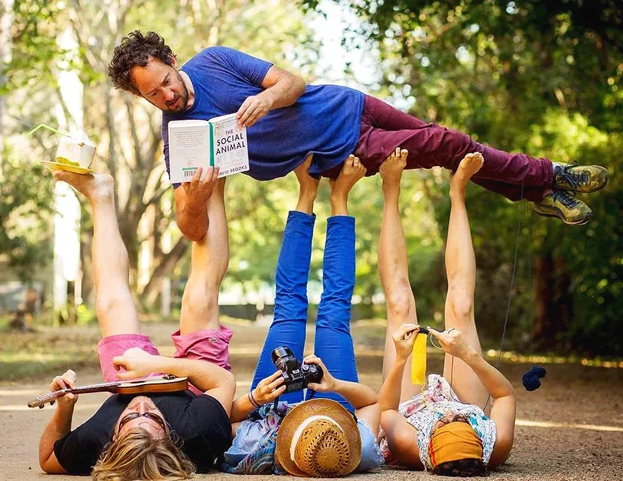 Three people lying on their backs on the ground, holding up a man who is lying horizontally above them. The man is reading a book, and the scene is set outdoors with trees in the background.