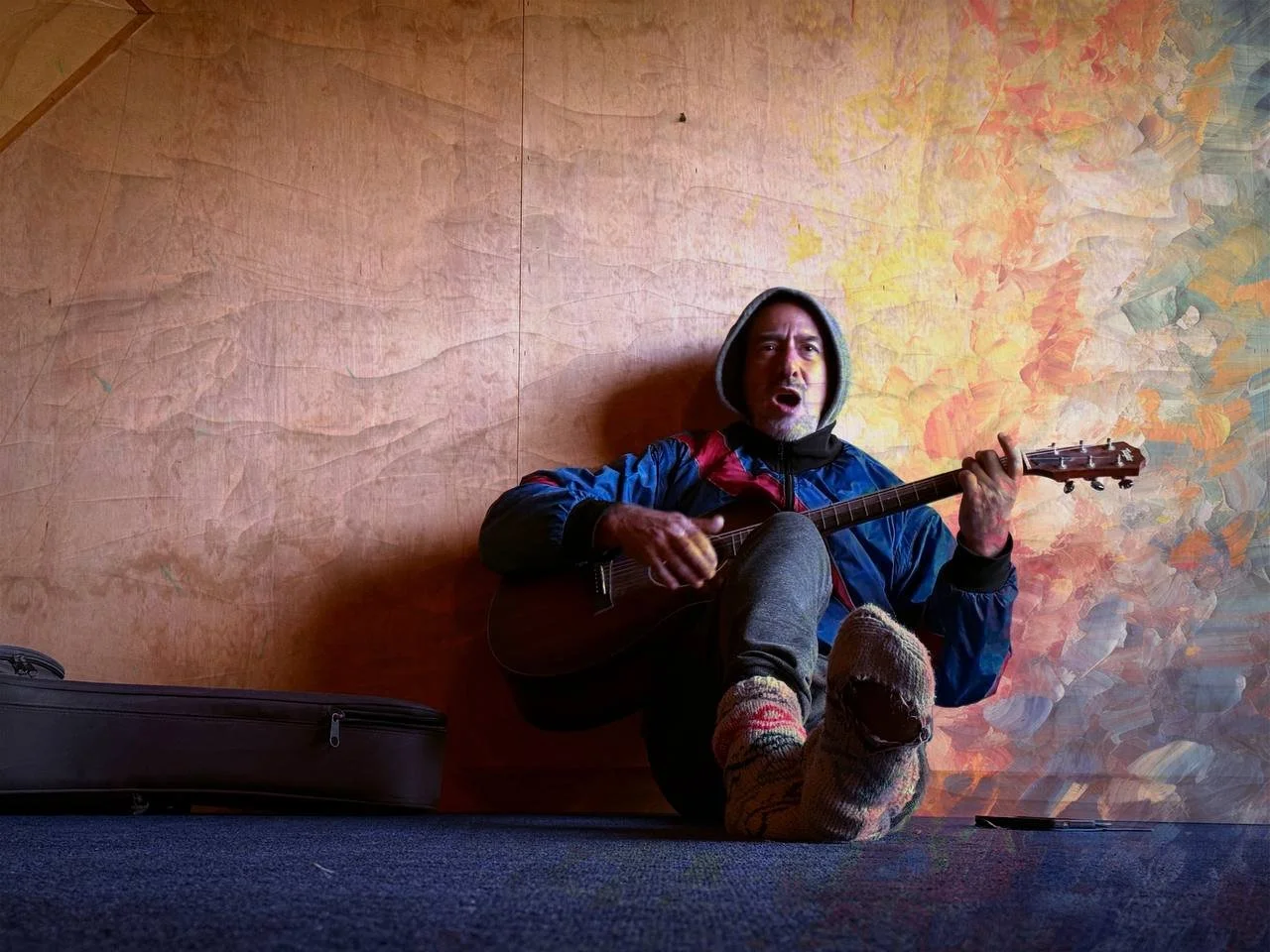 Jonno Katz sitting on the floor with his back against a wooden wall, playing an acoustic guitar. He's wearing a blue jacket, gray pants, and colorful socks, with his hood up and an expressive facial expression. wall is bland to coloured.