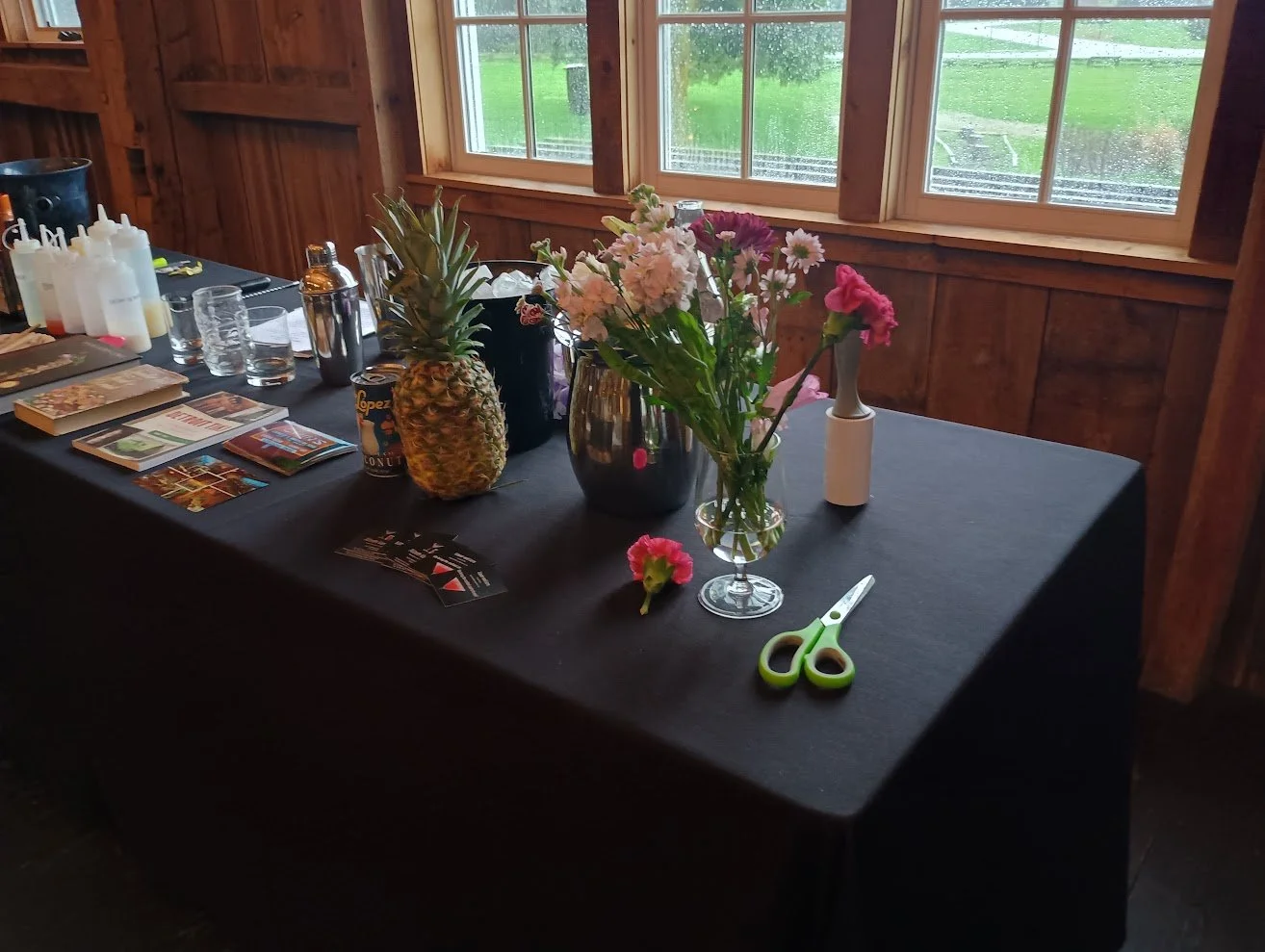 A black table with assorted vases holding flowers, a pineapple, scissors, and various items in a rustic wooden room with windows showing rain outside.